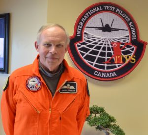 Geoff Connolly in orange flight suit in front of ITPS crest on wall.