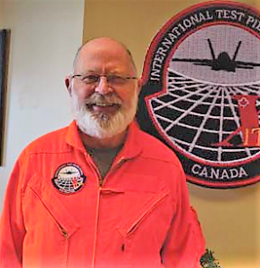 John Turner wearing an orange flight suit in front of ITPS crest on the wall.