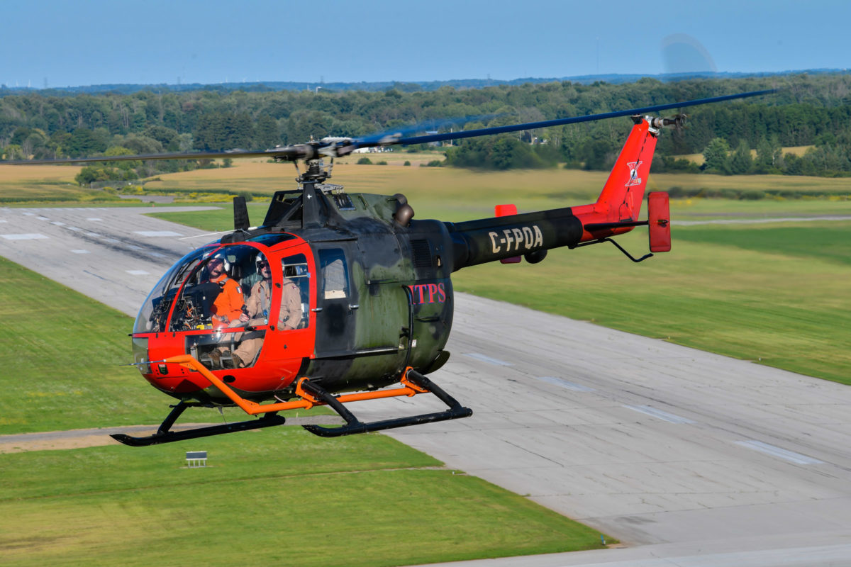 Green and red MBB Bo105 helicopter flying over the runway at CYXU