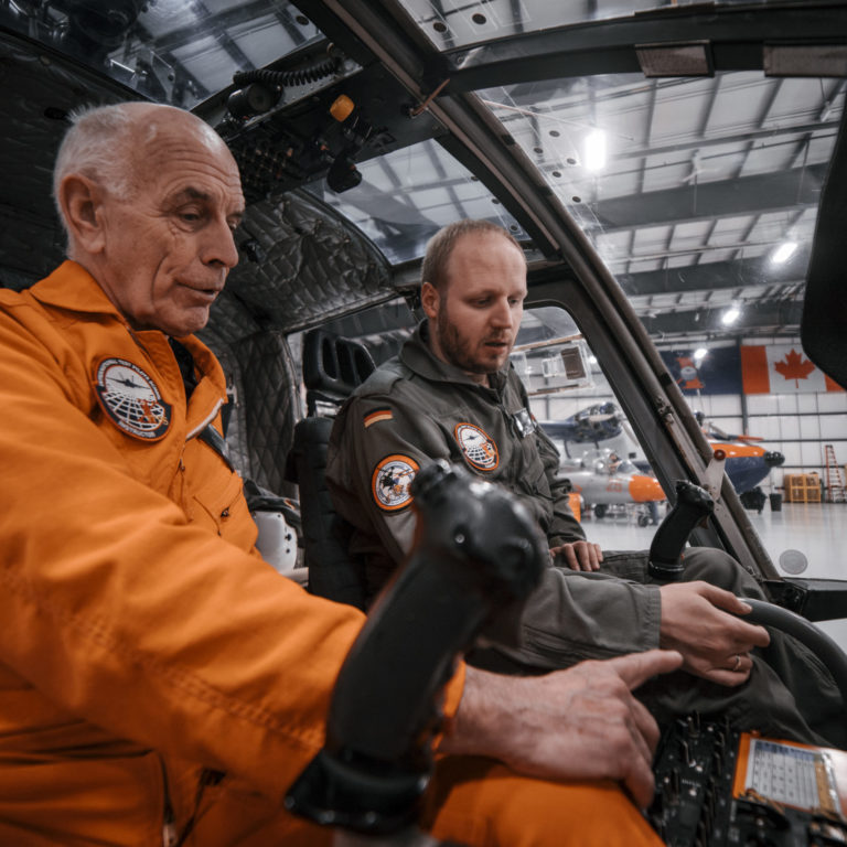An instructor wearing an orange jumpsuit and student wearing a green jumpsuit in the cockpit of an aircraft.