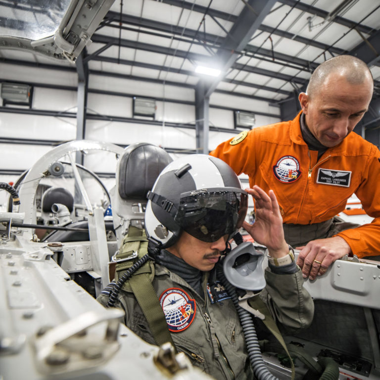 An instructor wearing an orange jumpsuit leaning over the edge of an aircraft and a student wearing a green jumpsuit seated in the cockpit with a mask, helmet and safety glasses.
